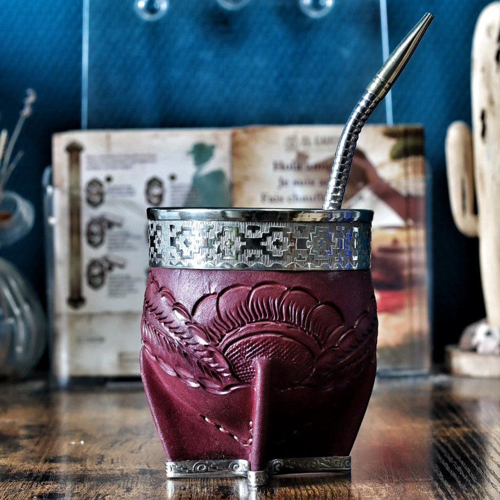 Calebasse à maté "Imperial" en cuir bordeaux labré avec une virole lisse en maillechort (alpaca). La bande de cuir centrale (le fleje) présente des détails de motifs Pampa gravés. Le récipient est posé sur une table en bois avec sa bombilla en acier.
