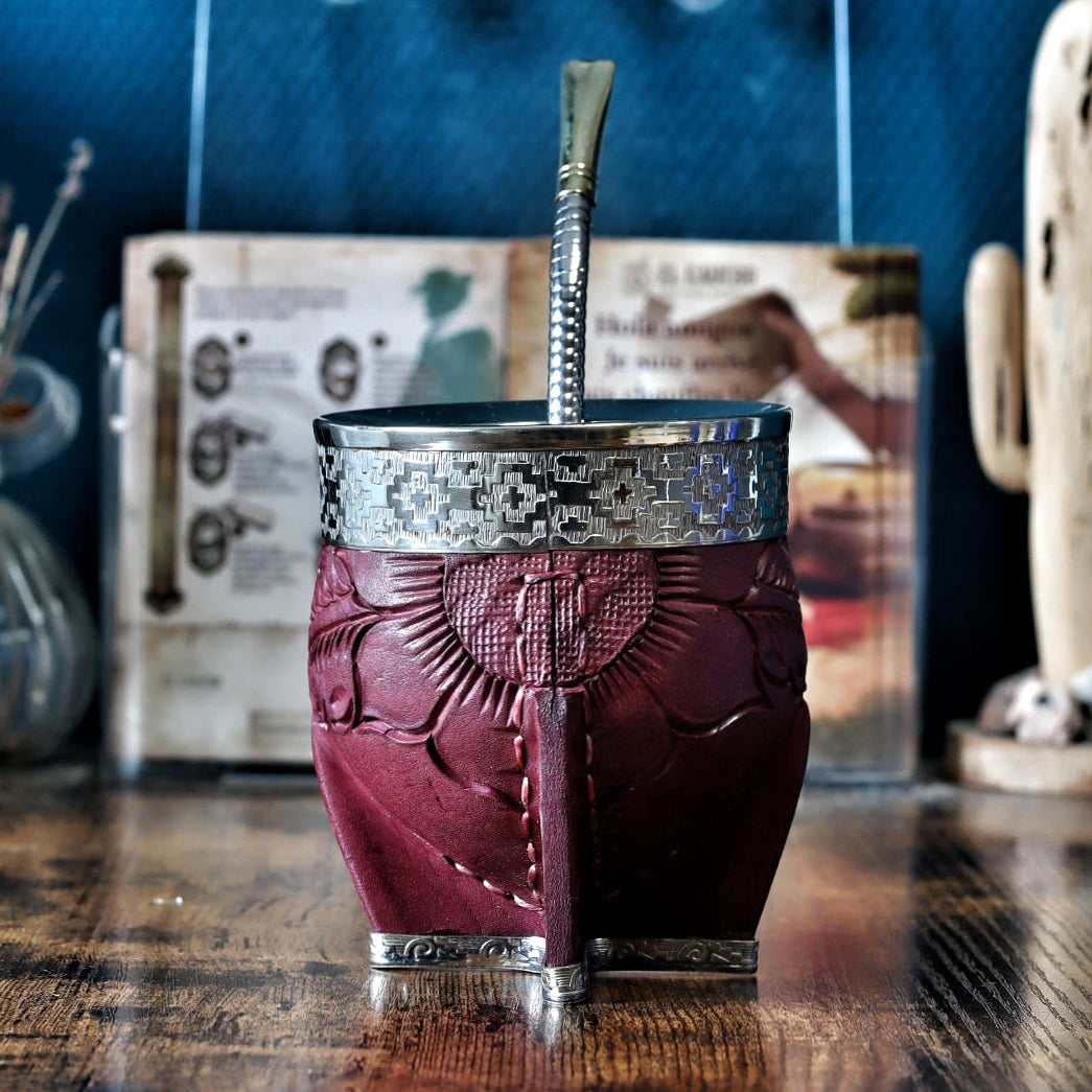 Calebasse à maté "Imperial" en cuir bordeaux labré avec une virole lisse en maillechort (alpaca). La bande de cuir centrale (le fleje) présente des détails de motifs Pampa gravés. Le récipient est posé sur une table en bois avec sa bombilla en acier.