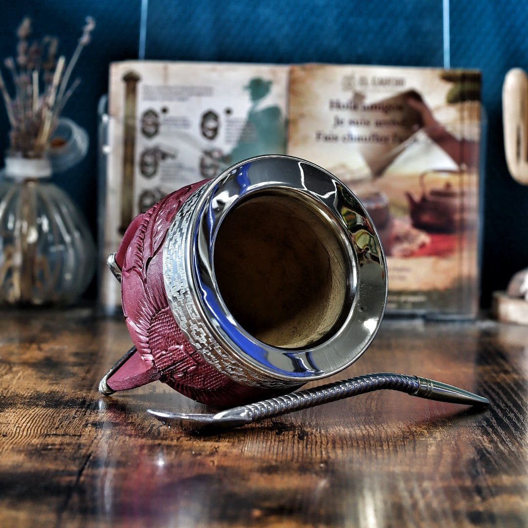Vue plongeante du maté Imperial bordeaux incliné, révélant l'intérieur en courge naturelle polie et l'éclat de la virole lisse en maillechort (alpaca). La bombilla en acier repose sur la table en bois au premier plan, devant les motifs Pampa de la bande de cuir.