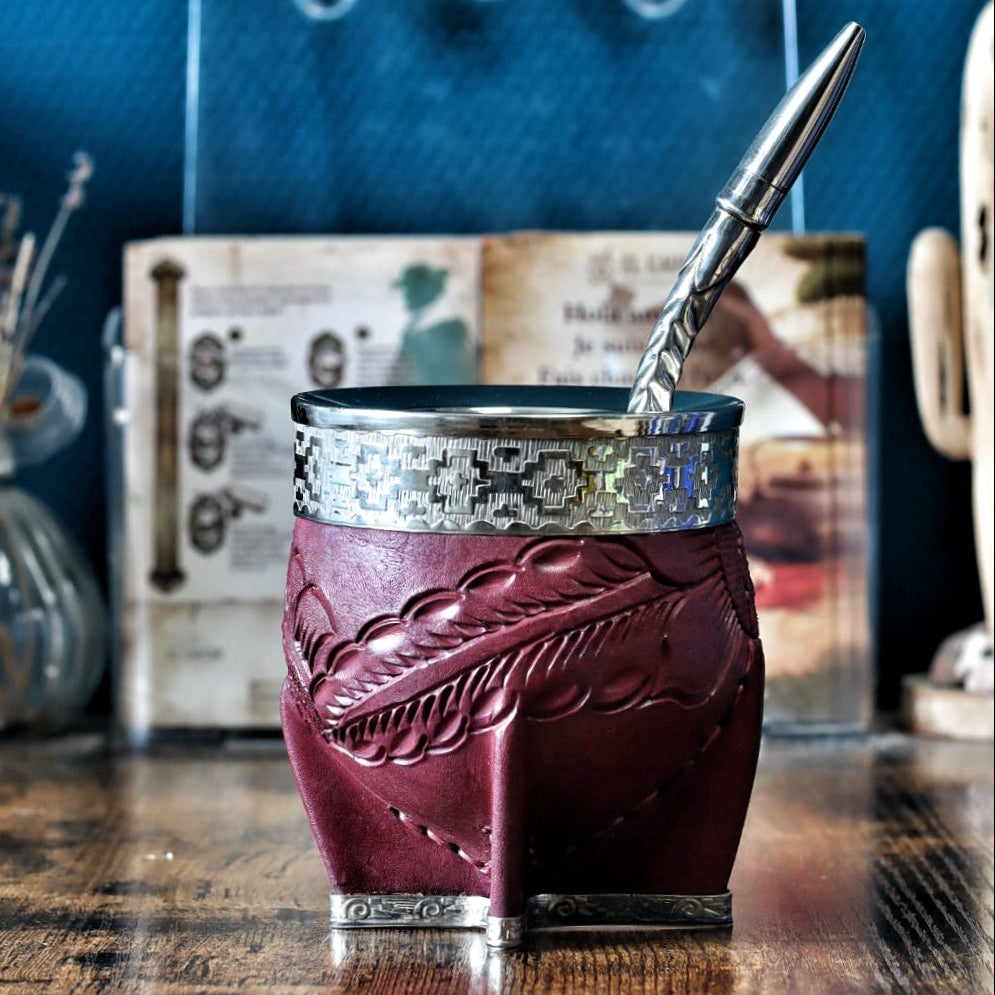 Calebasse à maté "Imperial" en cuir bordeaux labré avec une virole lisse en maillechort (alpaca). La bande de cuir centrale (le fleje) présente des détails de motifs Pampa gravés. Le récipient est posé sur une table en bois avec sa bombilla en acier.