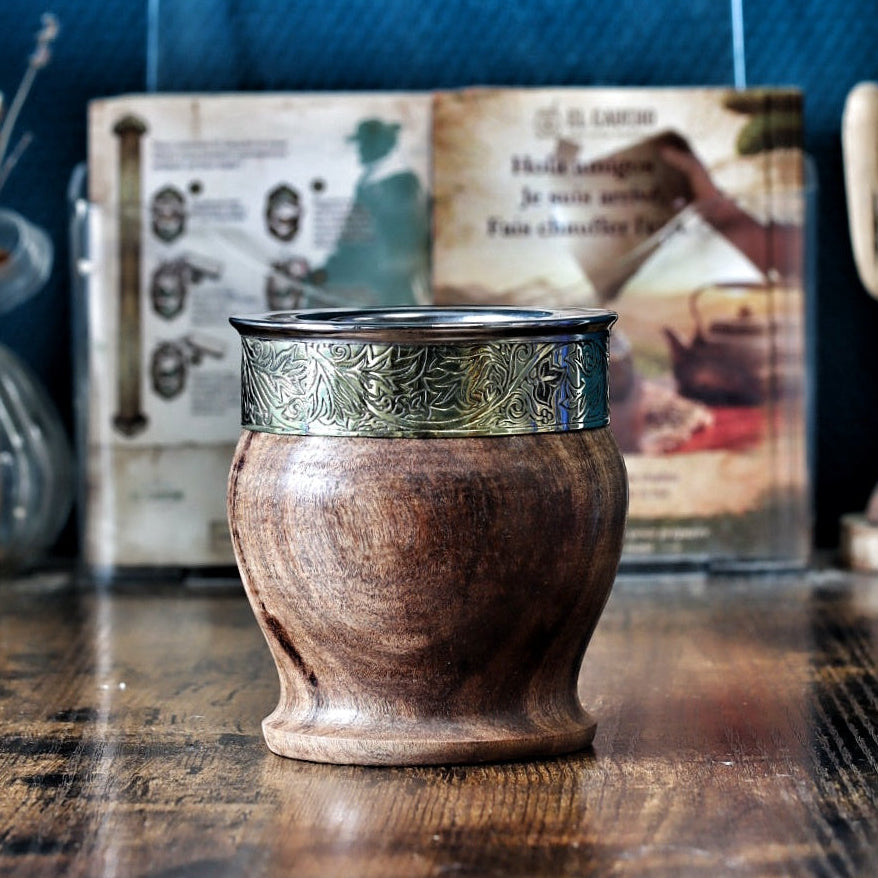 Vue détaillée d'un maté Impérial tonneau en bois de caroubier, surmonté d'une virole lisse en alpaga et décoré d'un "fleje" métallique finement ciselé avec de riches motifs floraux (feuilles et fleurs) pour une touche artisanale élégante.