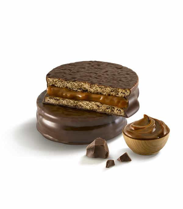 Alfajor Havanna au chocolat coupé montrant les deux biscuits moelleux garnis de dulce de leche crémeux, enrobé de chocolat mi-amer, avec morceaux de chocolat et cuillère de dulce de leche