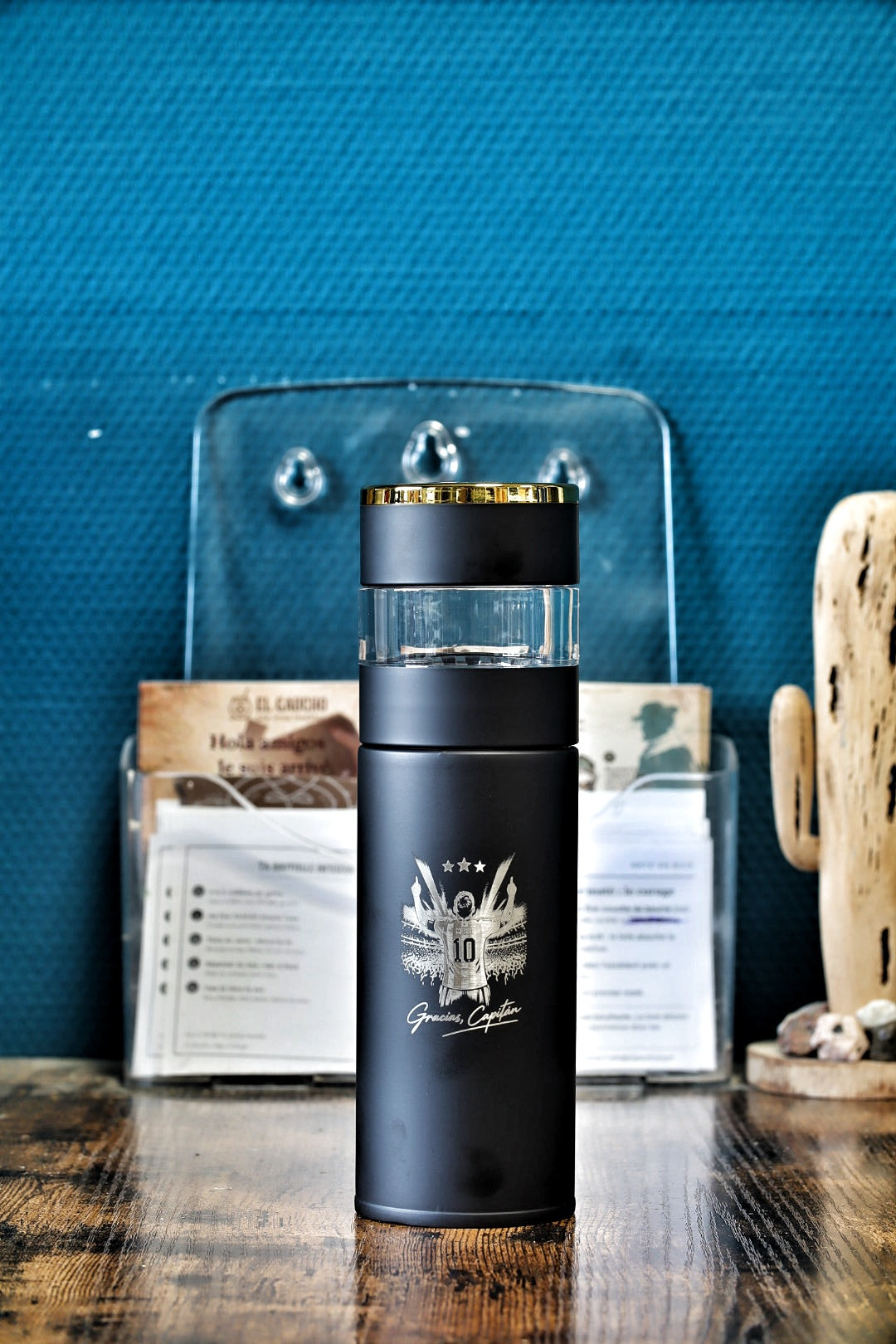 Bouteille infuseur noire au couvercle doré gravée « Gracias, Capitán » posée sur une table en bois