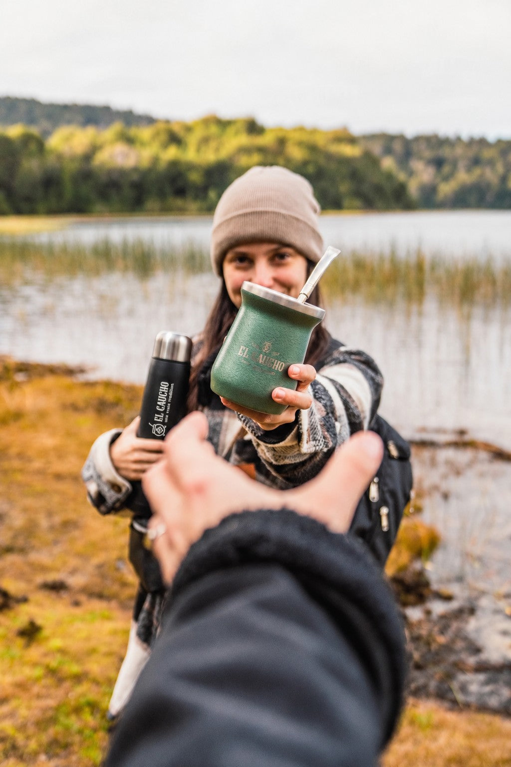 Au bord d'un lac, une matera tend son maté El Gaucho vert et garde le Mate Listo en mode thermos dans l'autre main