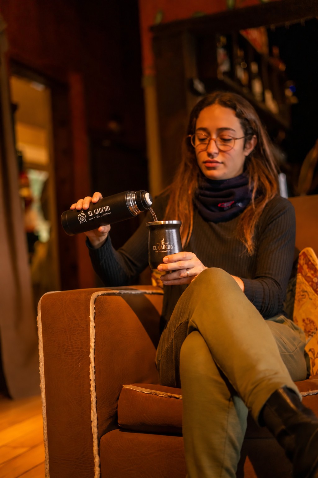 Assise dans un fauteuil, une matera se sert de l'eau avec le Mate Listo El Gaucho dans son maté noir